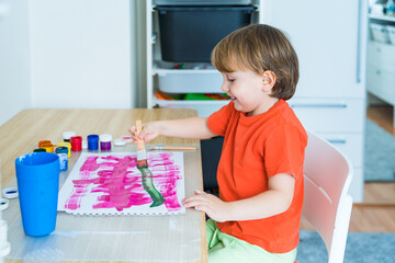 Little boy sitting at the desk and drawing colorful picture with paint and brushes. Child education at home during self isolation and lockdown. Concept of art and creativity of children.