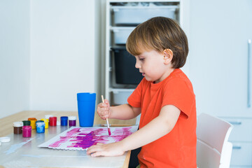 Little boy sitting at the desk and drawing colorful picture with paint and brushes. Child education at home during self isolation and lockdown. the child is holding the brush incorrectly