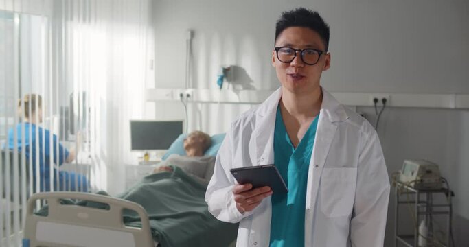 Happy smiling asian doctor in uniform holding tablet pc and talking at hospital ward
