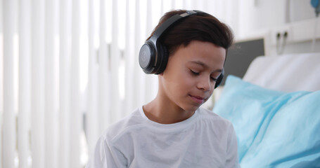 Smiling preteen african boy listening to music in headphone sitting in hospital bed