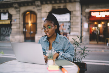 Dark skinned female sitting at street cafe with laptop computer doing remote job, youthful IT developer in eyewear typing and searching information from social networks during freelance work