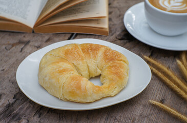 A croissant on a white ceramic plate and hot coffee on a wooden table.