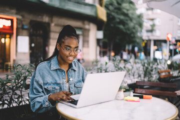 Obraz premium Dark skinned woman in glasses typing on laptop computer share content in blog browsing web pages and information,African American female freelancer doing remote job using wifi internet at cafe terrace