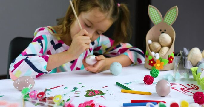 Happy little girl painting, drawing with brush eggs at home. Child preparing for Easter, having fun and celebrating feast. Happy Easter, DIY