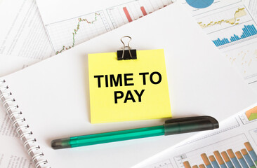 A yellow sticker with text Time To Pay is in a Notepad with a green pen financial charts and documents