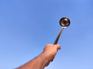 South Indian man hand holding silver laddle . A type of spoon used for soup, stew, or other foods. Cloud background.