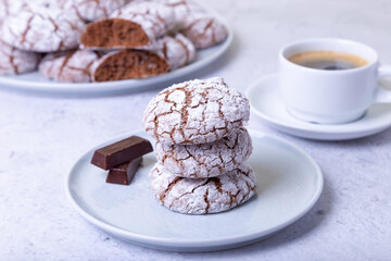 Chocolate cookies with cracks, strewed with icing sugar. Homemade baking. Three cookies in a stack. Background: a cup of coffee and a plate with cookies. Selective focus, close-up.
