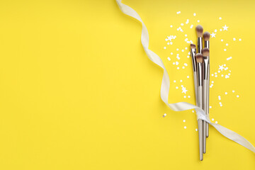 Different makeup brushes, ribbon and shiny confetti on yellow background, flat lay. Space for text