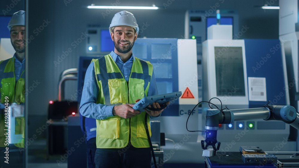 Poster Portrait of Professional Engineer Wearing Safety Vest and Hardhat, Uses Industrial Digital Tablet to Program, Manipulates Robot Arm, Looks at Camera and Smiles. Factory Workshop with CNC Machinery