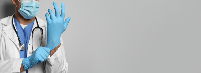 Doctor in protective mask putting on medical gloves against light grey background, closeup. Banner...