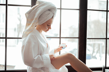 Young smiling blonde beautiful woman in the white bathrobe spread cream to her legs skin
