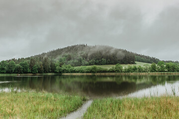 Fototapeta na wymiar Panoramic view of a lake in front of misty forest. Foggy morning outdoors.Beautiful autumn spring landscape with hill and water reflection.Misty forest scenery.Trees reflected in water.Calm atmosphere