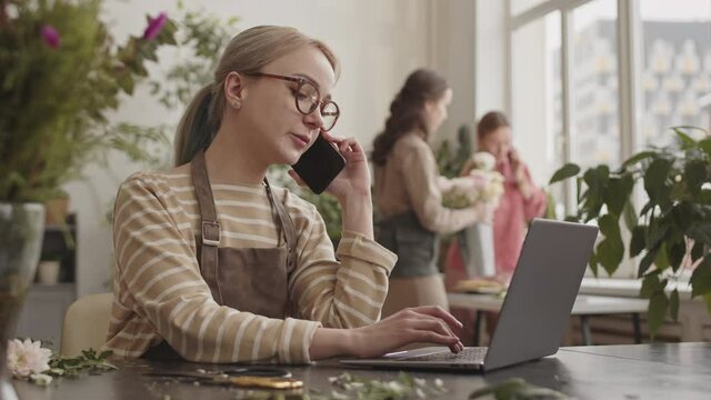 Medium close-up of blonde Caucasian woman wearing glasses, sitting by desk in flower shop, using laptop computer and talking on mobile phone, blurred colleagues working on background