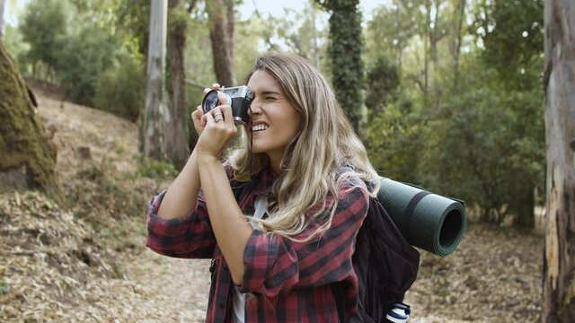 Happy backpacker girl with photo camera taking pictures of forest landscape, while walking on path, enjoying outdoor leisure time and healthy recreation. Adventure travel concept