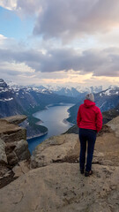 A girl wearing pink jacket stands next to the hanging rock formation Trolltunga with a view on Ringedalsvatnet lake, Norway at the sunrise. Slopes are partially covered with snow Freedom and happiness