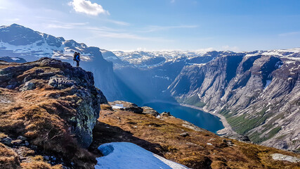 A young man wearing blue jacket and huge hiking backpack stands on a tall rock, admiring the view...