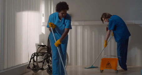 Diverse janitors washing floor in hospital room with empty wheelchair