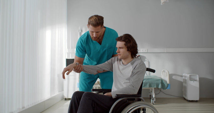 Nurse lifting arm of young paralyzed man sitting in wheelchair