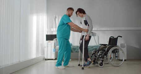 Smiling doctor helping young man patient with crutches.