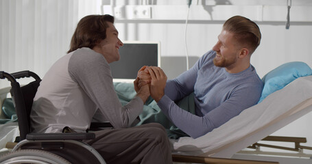 Disabled young man in wheelchair holding hands of sick boyfriend lying in hospital bed