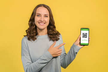 Smiling young woman points to online covid passport on the smartphone, isolated on yellow, lady shows an e-document confirmatory vaccination, necessary app for travel during and after pandemic period