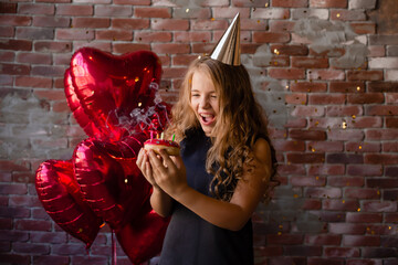 Happy little girl makes a wish and blows out candles on a cake. brick background, balloons. Space...