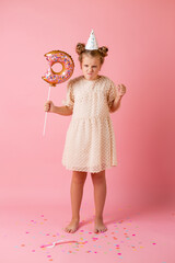 A happy little girl in a birthday cap holds a doughnut-shaped balloon on a pink background in the studio, space for text