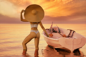 Young beautiful sexy fit brunette  model pose on the beach with huge beach hat at caramel milky orange sunset next to fishing boat