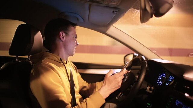 The taxi driver has lost his address while driving. An expression of misunderstanding, looking around. He looks at the navigation application of his phone in the night city.