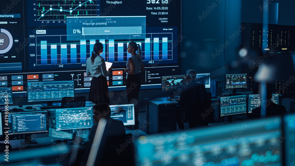 Poster Two Software Engineers Talking Next to a Big Screen in a Modern Monitoring Office with Live Analysis Feed with Charts. Monitoring Room Big Data Scientists and Managers Sit in Front of Computers.
