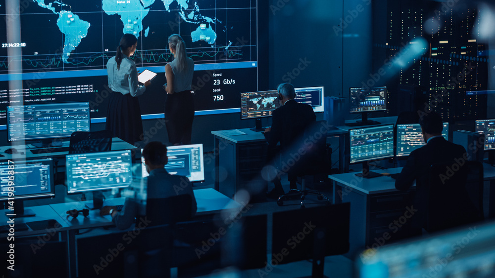 Poster Two Software Engineers Talking Next to a Big Screen in a Modern Monitoring Office with Live Analysis Feed with Charts. Monitoring Room Big Data Scientists and Managers Sit in Front of Computers.