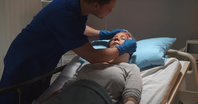 Mae nurse washing wound of unconscious aged female patient lying in hospital bed