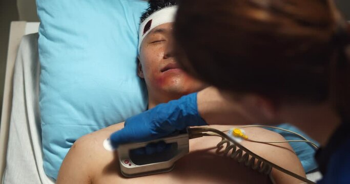 Close up of doctor using defibrillator on injured asian male patient in ward at hospital.