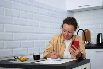 Happy young woman in headphones using smartphone sitting at the table in kitchen at home,writing notes.Smiling curly female student watching online webinar,learning language,listening audio course.