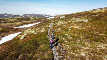 A couple hiking through the highlands of Eidfjors, Norway. Both of them carry big backpacks. Hike into the wilderness. Snow covers some parts of the slopes. Dry grass around the trail. Rough hike.