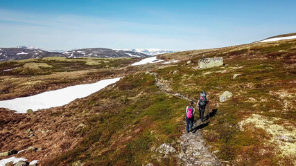 Fototapeta premium A couple hiking through the highlands of Eidfjors, Norway. Both of them carry big backpacks. Hike into the wilderness. Snow covers some parts of the slopes. Dry grass around the trail. Rough hike.