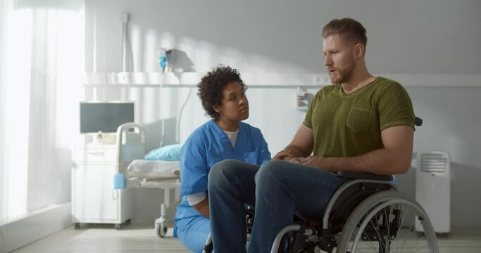 African female nurse supporting and cheering upset male patient in wheelchair
