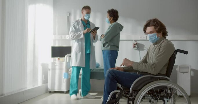 Sad man in surgical mask sitting on wheelchair in hospital room while girlfriend talking to doctor
