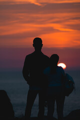 Young couple in love admiring a beautiful red and orange sunset in a famous sunset point in Sardinia, Italy. Romantic moment for the two, framed by the sun an the scenic sky.