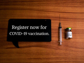 Top view of a syringe, vial covid-19 vaccine and wooden craft written a "Register now for COVID-19 vaccination" isolated on a wooden background. Covid-19 vaccination concept.