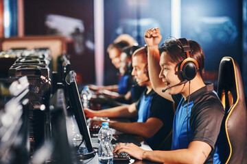 Side view of excited young asian guy, male cyber sport gamer looking at PC screen and raising hand up while - obrazy, fototapety, plakaty