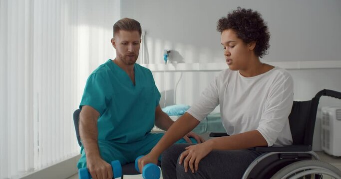 Doctor helping patient in wheelchair with dumbbells doing rehabilitation exercise in medical center