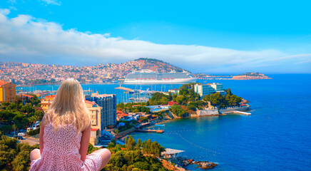 Fototapeta na wymiar Lone young blonde girl sitting on a rock looking at the sea - Cruise ships at port of Kusadasi - Aydin, Turkey