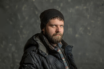 A brutal man with a beard in a knitted hat, plaid shirt and jacket looks at the camera, dark background. hipster, programmer, woodcutter, bully