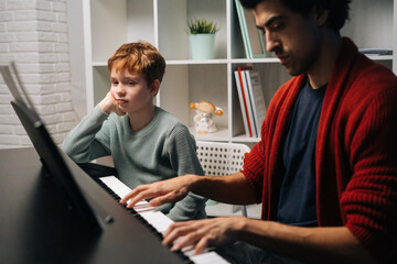 Bored redhead child student boy listening his teacher play the piano during lesson. Loving dad teaching talented son to play musical instrument in living room. Concept of music education.