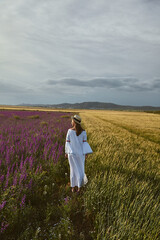 Fototapeta premium Young woman in hat posing in a wheat field with purple Consolida flowers on a summer day