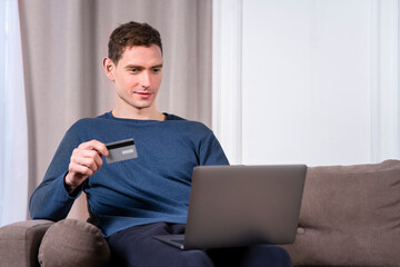 Happy cheerful young guy, handsome man with credit bank card paying online in shop, enter information, shopping, ordering in internet store, doing payment on laptop computer, smiling at home at couch