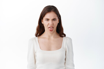 Obraz premium Teen girl showing tongue from dislike and aversion, something taste bad, frowning and complaining, staring displeased, standing against white background