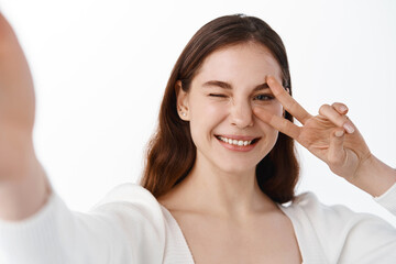 Attractive happy woman taking selfie with v-sign, stretch out hand to hold camera, making positive face, smile and peace gesture, standing over white background