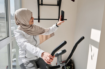 Attractive Muslim woman doing selfie exercising on a stationary bike. Cardio workout. Fitness at...